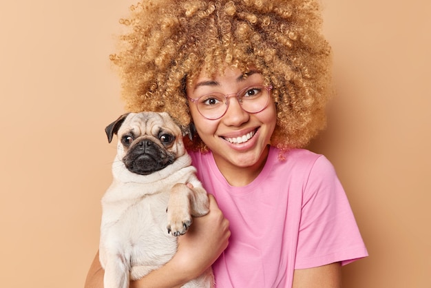 Porträt einer glücklichen Europäerin mit blonden, lockigen Haaren posiert mit Pug-Hund, der froh ist, dass Haustierbesitzer posiert, um ein unvergessliches Foto zu machen, das in einem rosafarbenen, lässigen T-Shirt gekleidet ist, das über beigem Hintergrund isoliert ist