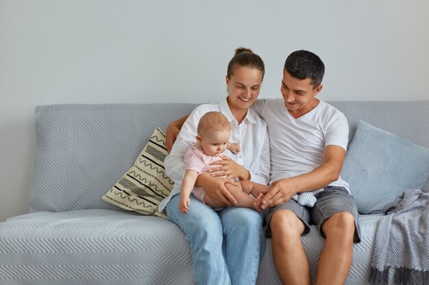 Porträt einer glücklichen Familie, die auf dem Sofa im Wohnzimmer sitzt, Menschen, die Freizeitkleidung tragen, Zeit mit ihrem Baby zu Hause verbringen, Elternschaft, Kindheit.