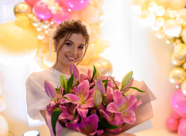 Porträt einer glücklichen Frau mit brünetter Frisur, gekleidet in ein trendiges Kleid, das ein großes Blumenarrangement mit rosa Lilien hält, die lächeln und während der Hochzeitsfeier im Restaurant in die Kamera schauen