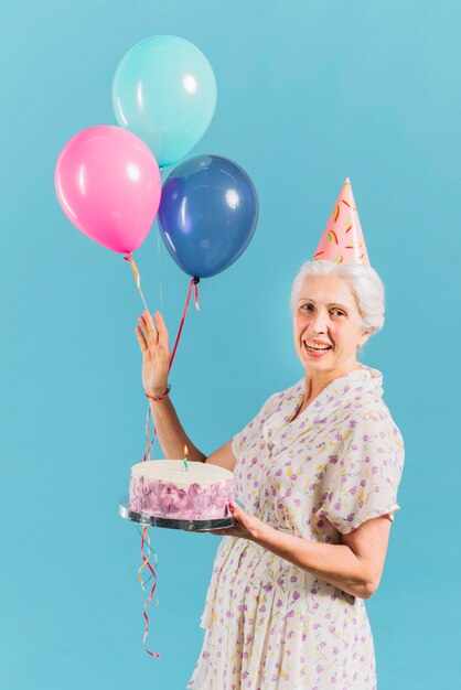 Porträt einer glücklichen Frau mit Geburtstagskuchen und -ballonen auf blauem Hintergrund
