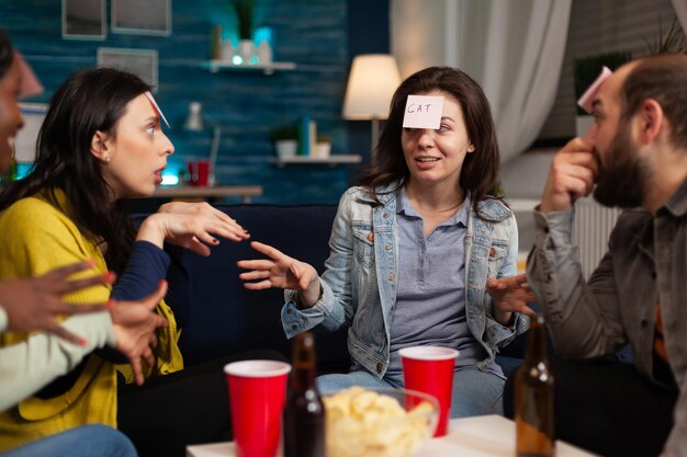 Porträt einer glücklichen Frau mit Haftnotizen auf der Stirn, die erraten, wer das Spiel genießt, Zeit mit Freunden zu verbringen, die während der Wochenendparty zu Hause zusammen sind. Freundschaftskonzept