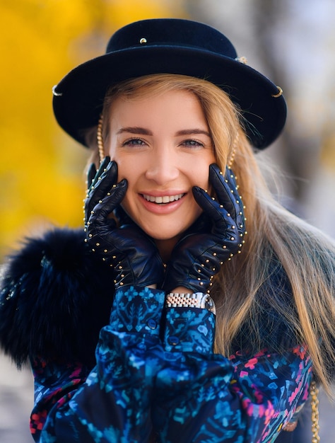 Porträt einer glücklichen Frau mit langen blonden Haaren, die eine Pelzweste mit schwarzem Hut und ein gemustertes Hemd trägt, die Hände in der Nähe des Gesichts hält, lächelt und in die Kamera schaut, während sie im Freien geht