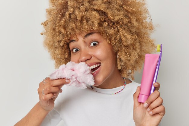 Porträt einer glücklichen Frau mit lockigem Haar isst Zuckerwatte, die für Ihre Zähne schädlich ist, hält eine Zahnpastatube und Zahnbürste hat einen frohen Ausdruck, der vor weißem Hintergrund posiert Mundhygiene und süße Speisen