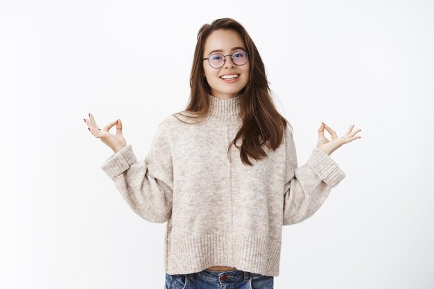 Porträt einer glücklichen, friedlichen, gutaussehenden Frau mit Brille und warmem Pullover, die ruhig bleibt, während sie meditiert und breit in die Kamera lächelt, die Hände mit Mudra-Zen-Kugeln hebt, die Yoga-Wissen über die graue Wand teilen