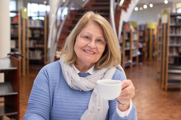 Kostenloses Foto porträt einer glücklichen gealterten frau, die in der bibliothek studiert. lächelnde kaukasische frau, die bücher liest, die sich auf computerkurse vorbereiten und sich bei einer tasse tee kurz ausruhen. selbstbildungskonzept für erwachsene