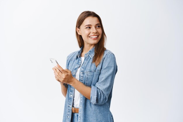 Porträt einer glücklichen, gutaussehenden Frau mit blonden Haaren, Blick direkt auf den Kopierbereich, der das Smartphone hält, das in sozialen Medien chattet, oder den weißen Hintergrund der mobilen Anwendung verwendet