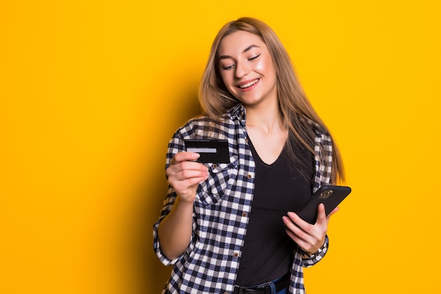 Porträt einer glücklichen jungen blonden Frau, die Plastikkreditkarte zeigt, während Handy verwendet, das über gelber Wand lokalisiert wird