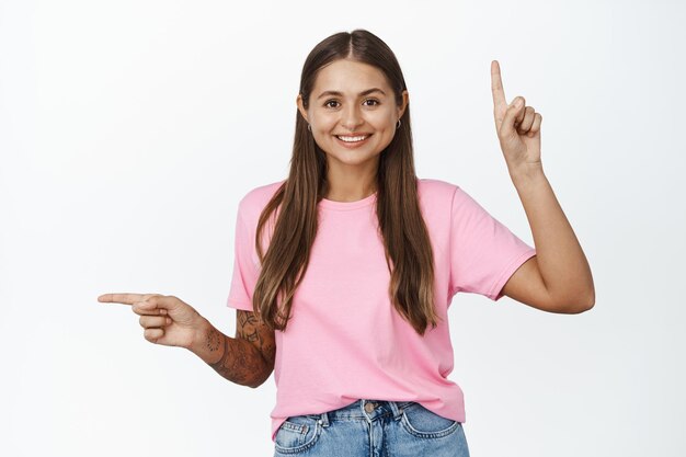 Porträt einer glücklichen jungen Frau, die lächelt, mit dem Finger auf zwei Seiten zeigt, nach links und oben zeigt und vor weißem Hintergrund steht.