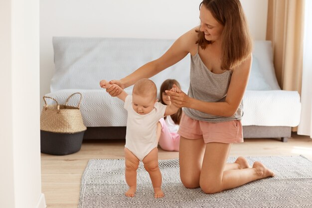 Porträt einer glücklichen Mutter, die auf den Knien auf dem Boden im Wohnzimmer steht und seinem Säugling beibringt, zu gehen, Kleinkindmädchen, das geht, glückliche Kindheit.