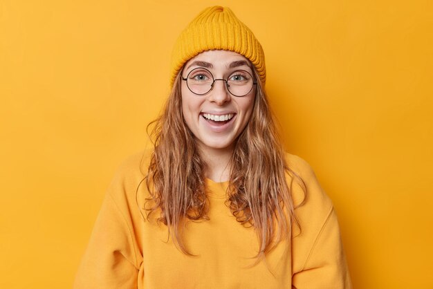 Porträt einer glücklichen, schönen Europäerin mit langen Haaren, blauen Augen, weißen, gleichmäßigen Zähnen, trägt einen runden, transparenten Brillenhut und ein Sweatshirt, die auf gelbem Hintergrund isoliert sind Positive Emotionen