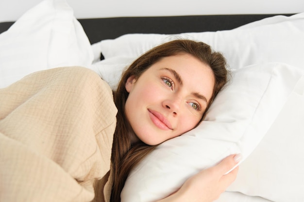 Porträt einer glücklichen, schönen jungen Frau, die im Schlafanzug im Bett liegt und ihr Kissen umarmt