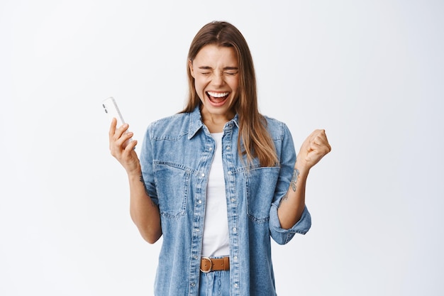 Porträt einer glücklichen schreienden Frau, die auf dem Smartphone gewinnt, das Handy hält und jubelt, den Sieg oder die Leistung im Internet feiert, weiß