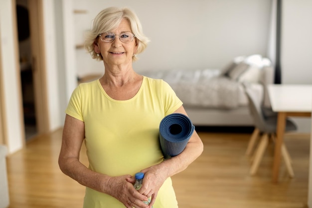 Porträt einer glücklichen Seniorin, die zu Hause eine Trainingsmatte und eine Flasche Wasser hält