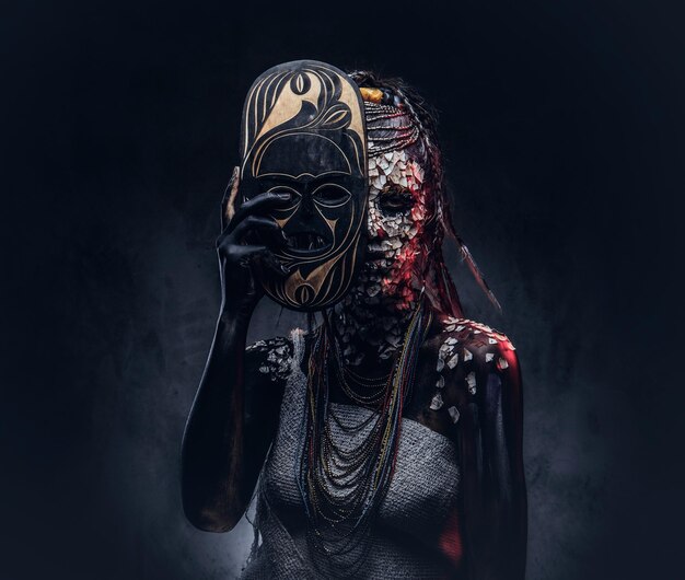 Porträt einer gruseligen afrikanischen Schamanin mit versteinerter, rissiger Haut und Dreadlocks, hält eine traditionelle Maske auf dunklem Hintergrund. Make-up-Konzept.