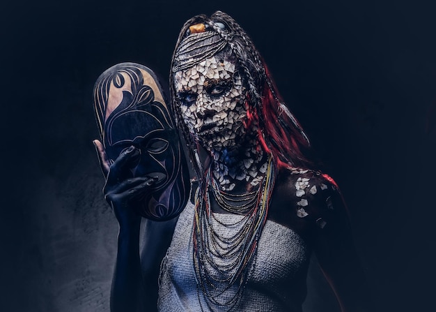Porträt einer gruseligen afrikanischen Schamanin mit versteinerter, rissiger Haut und Dreadlocks, hält eine traditionelle Maske auf dunklem Hintergrund. Make-up-Konzept.