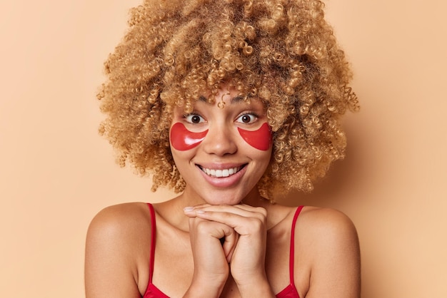 Porträt einer gut aussehenden europäerin mit lockigem buschigem haar lächelt angenehm und hält die hände unter dem kinn trägt rote hydrogelflecken unter den augen auf, die lässig isoliert über braunem hintergrund gekleidet sind