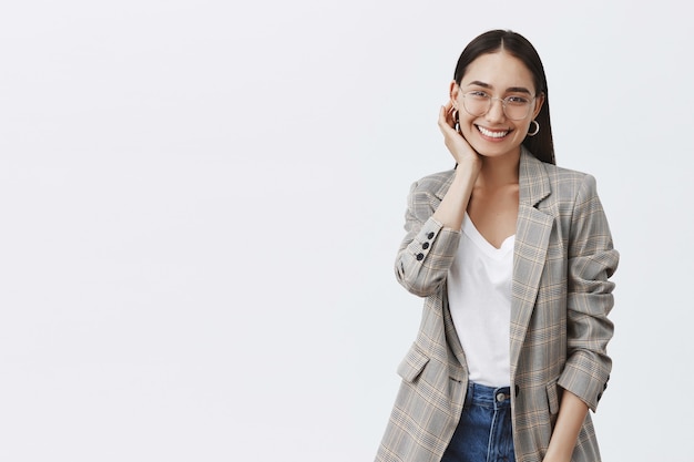 Porträt einer gut aussehenden stilvollen Frau in Brille und trendiger Jacke, die den Ohrring berührt, während sie über die graue Wand flirtet