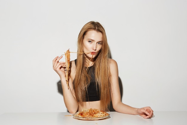 Porträt einer gutaussehenden jungen Frau mit langen hellen Haaren in schwarzem Oberteil, die Spaghetti mit Gabel isst und entspannt in die Kamera schaut