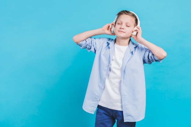 Porträt einer hörenden Musik des Jungen auf weißem Kopfhörer gegen blauen Hintergrund