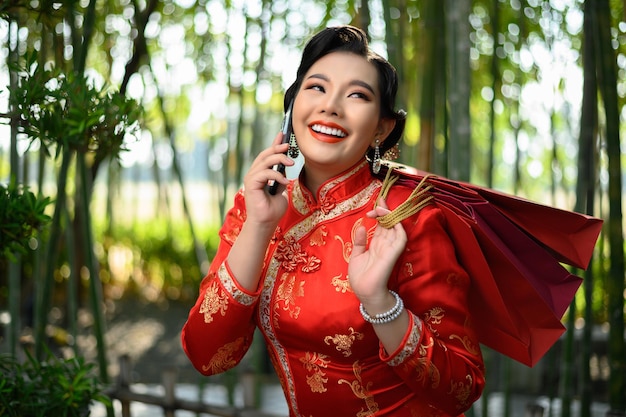 Porträt einer hübschen asiatischen Frau in einem chinesischen Cheongsam, die Einkaufstüten hält und mit dem Handy mit Glück im Bambuswald spricht