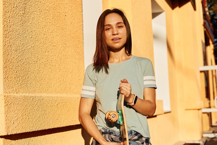 Kostenlose Foto porträt einer hübschen brünetten frau mit t-shirt, die mit skateboard in den händen steht und die kamera mit ruhigem gesichtsausdruck betrachtet und im freien gegen gelbes gebäude posiert.