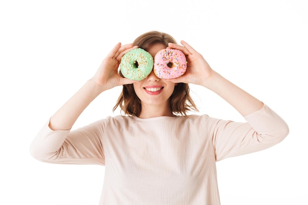 Porträt einer hübschen Dame in rosafarbenem Kleid, die steht und ihre Augen mit Donuts auf weißem Hintergrund bedeckt