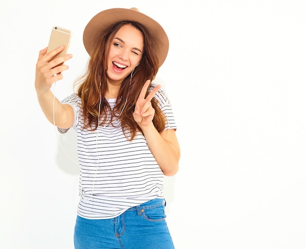 Porträt einer hübschen frau im sommerhippie kleidet das nehmen eines selfie, das auf weißer wand lokalisiert wird. friedenszeichen blinzeln und zeigend