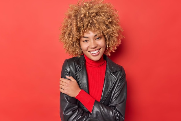 Porträt einer hübschen, fröhlichen Frau mit blonden Locken, die zahnig lächelt und eine modische Lederjacke trägt, fühlt sich zufrieden, nachdem sie ein neues Outfit einzeln auf leuchtend rotem Hintergrund gekauft hat. Positive Gefühle