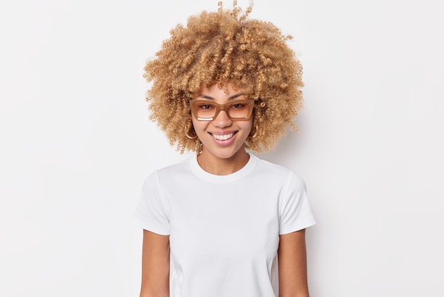 Porträt einer hübschen jungen Frau mit lockigem Haar, die glücklich lächelt, trägt ein lässiges Basic-T-Shirt mit Brille und hat einen fröhlichen Ausdruck.