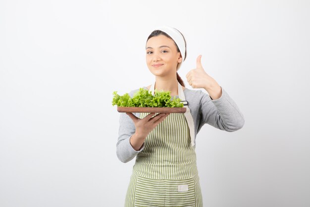 Porträt einer hübschen süßen Frau mit einem Holzbrett aus frischem Salat, das einen Daumen nach oben zeigt