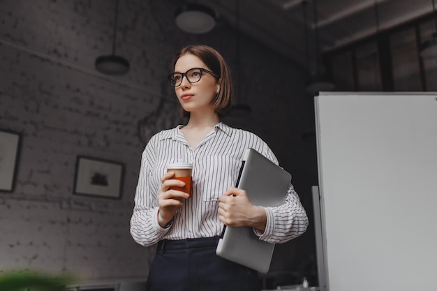 Porträt einer intelligenten Frau mit Brille, die eine Tasse Tee hält Mitarbeiterin in stilvollem Outfit posiert mit Laptop