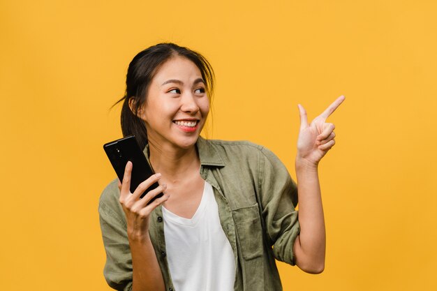 Porträt einer jungen asiatischen Dame, die Handy mit fröhlichem Ausdruck benutzt, etwas Erstaunliches an leeren Stellen in Freizeitkleidung zeigt und isoliert über gelber Wand steht. Gesichtsausdruck Konzept.