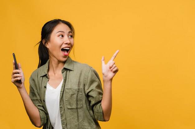 Porträt einer jungen asiatischen Dame, die Handy mit fröhlichem Ausdruck benutzt, etwas Erstaunliches an leeren Stellen in Freizeitkleidung zeigt und isoliert über gelber Wand steht. Gesichtsausdruck Konzept.