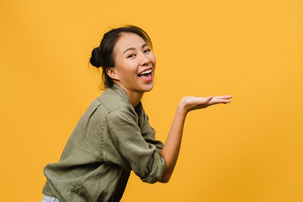 Porträt einer jungen asiatischen Dame, die mit fröhlichem Ausdruck lächelt, zeigt etwas Erstaunliches im leeren Raum in legerem Tuch, das über gelber Wand isoliert ist. Gesichtsausdruck Konzept.