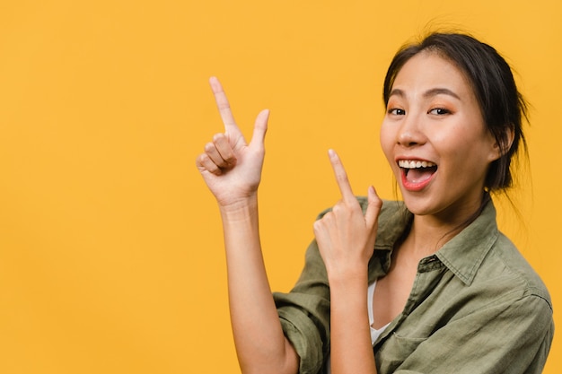 Porträt einer jungen asiatischen Dame, die mit fröhlichem Ausdruck lächelt, zeigt etwas Erstaunliches im leeren Raum in legerem Tuch, das über gelber Wand isoliert ist. Gesichtsausdruck Konzept.