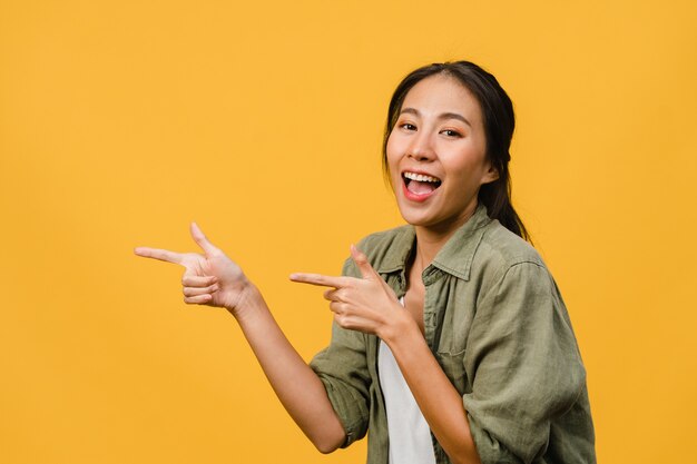 Porträt einer jungen asiatischen Dame, die mit fröhlichem Ausdruck lächelt, zeigt etwas Erstaunliches im leeren Raum in legerem Tuch, das über gelber Wand isoliert ist. Gesichtsausdruck Konzept.