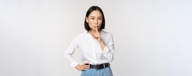 Porträt einer jungen asiatischen erwachsenen Frau, die zum Schweigen bringt, sagen Sie, hush, shh, drücken Sie den Finger auf die Lippen, um das Geheimnis zu teilen, sprechen Sie keine Tabu-Geste, die über weißem Hintergrund steht