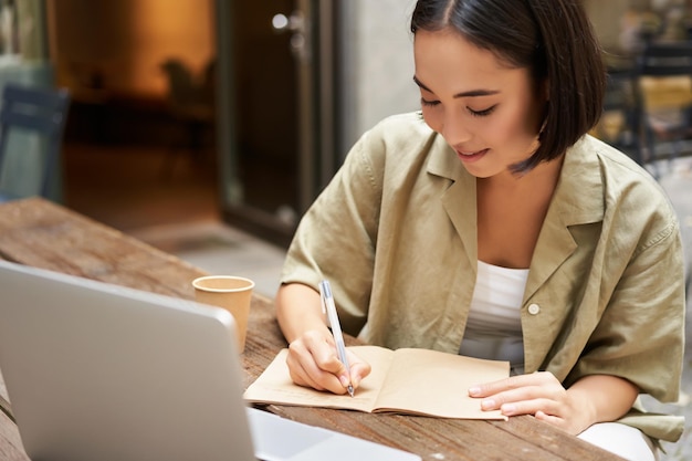 Porträt einer jungen asiatischen Frau, die am Laptop arbeitet und sich Notizen macht, während sie an einem Arbeitstreffen für Online-Unterricht teilnimmt