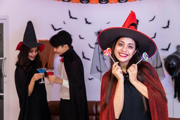 Porträt einer jungen asiatischen Frau, die im Halloween-Kostüm als Hexe auf einer Party lächelt und Süßigkeiten hält, die in die Kamera schauen
