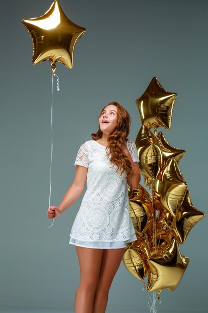 Porträt einer jungen attraktiven Frau in einem weißen Kleid, die viele goldene Luftballons hält, auf hellem Hintergrund