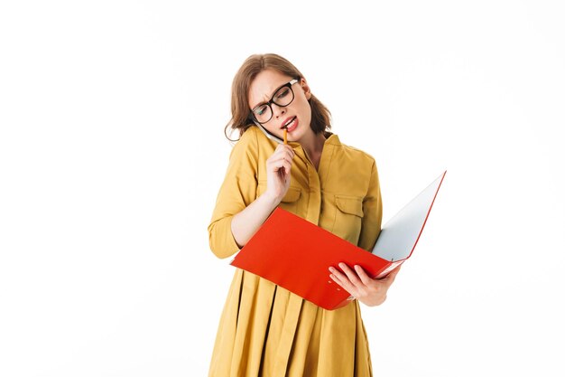 Porträt einer jungen beschäftigten Dame mit Brille, die auf ihrem Handy spricht, während sie nachdenklich mit offenem rotem Ordner und Bleistift in der Hand auf weißem Hintergrund zur Seite schaut