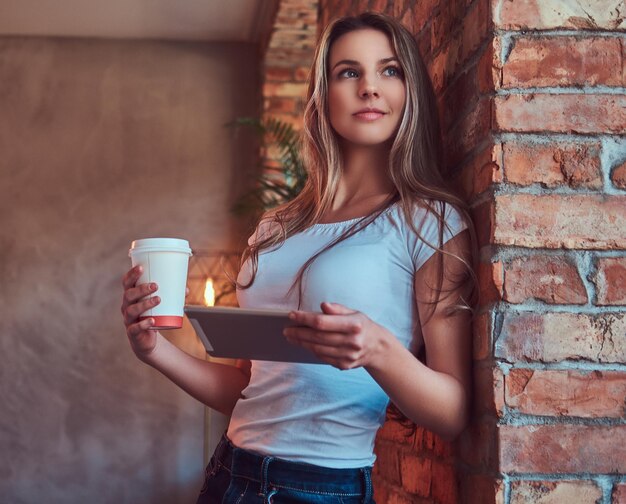 Porträt einer jungen blonden Frau hält eine Tasse Kaffee zum Mitnehmen und ein digitales Tablet, während sie sich in einem Raum mit Loft-Interieur an eine Ziegelwand lehnt.