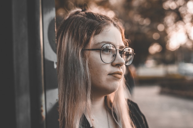 Kostenloses Foto porträt einer jungen blonden frau mit brille, die an eine wand lehnt und sanft lächelt