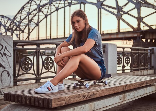 Porträt einer jungen brünetten Frau in T-Shirt und Shorts, die bei Sonnenuntergang auf einem Skateboard auf dem Hintergrund der alten Brücke sitzt.