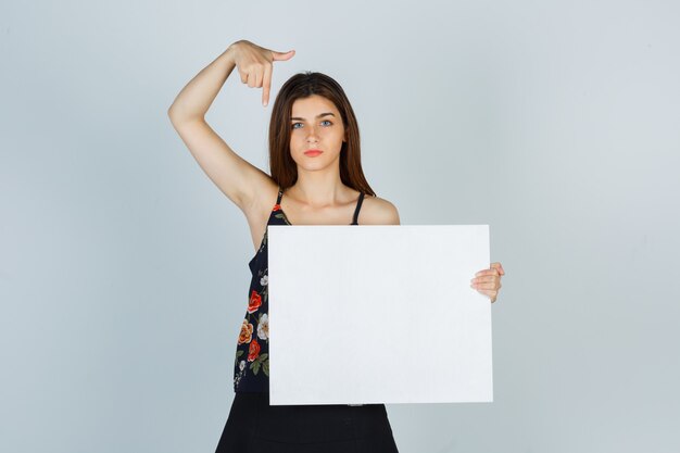 Porträt einer jungen Dame, die auf leere Leinwand in Bluse, Rock zeigt und ernsthafte Vorderansicht sieht