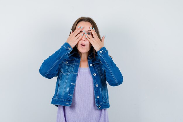 Porträt einer jungen Dame, die durch die Finger in T-Shirt, Jacke schaut und neugierige Vorderansicht schaut