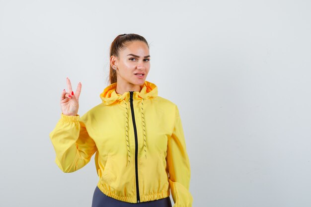 Porträt einer jungen Dame, die Siegesgeste in gelber Jacke zeigt und selbstbewusste Vorderansicht sieht