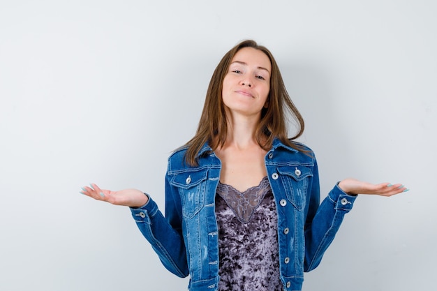 Porträt einer jungen Dame, die Waagengeste in Bluse, Jeansjacke und fröhlicher Vorderansicht macht