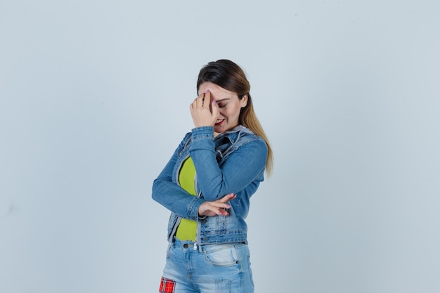 Porträt einer jungen dame mit der hand auf dem gesicht, während sie in jeans-outfit lächelt und sich schämt vorderansicht