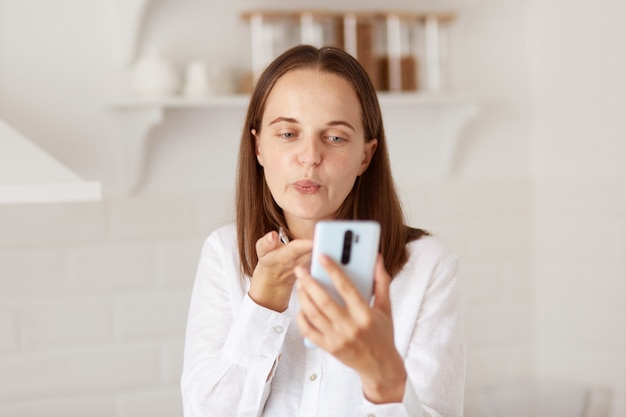Porträt einer jungen erwachsenen, glücklichen Frau, die Smartphone für Videoanrufe in der Küche zu Hause verwendet und Luftküsse für Anhänger sendet, während sie Livestream sendet.